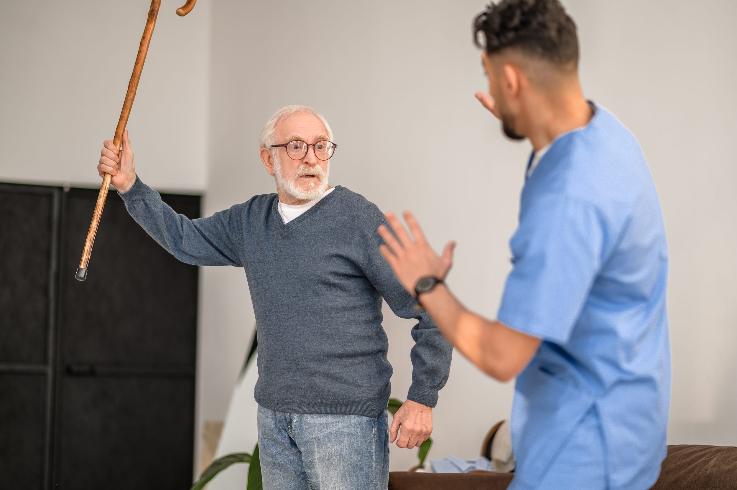 Code Gray Irritated gray-haired old man brandishing the walking stick at his in-home caregiver standing before him