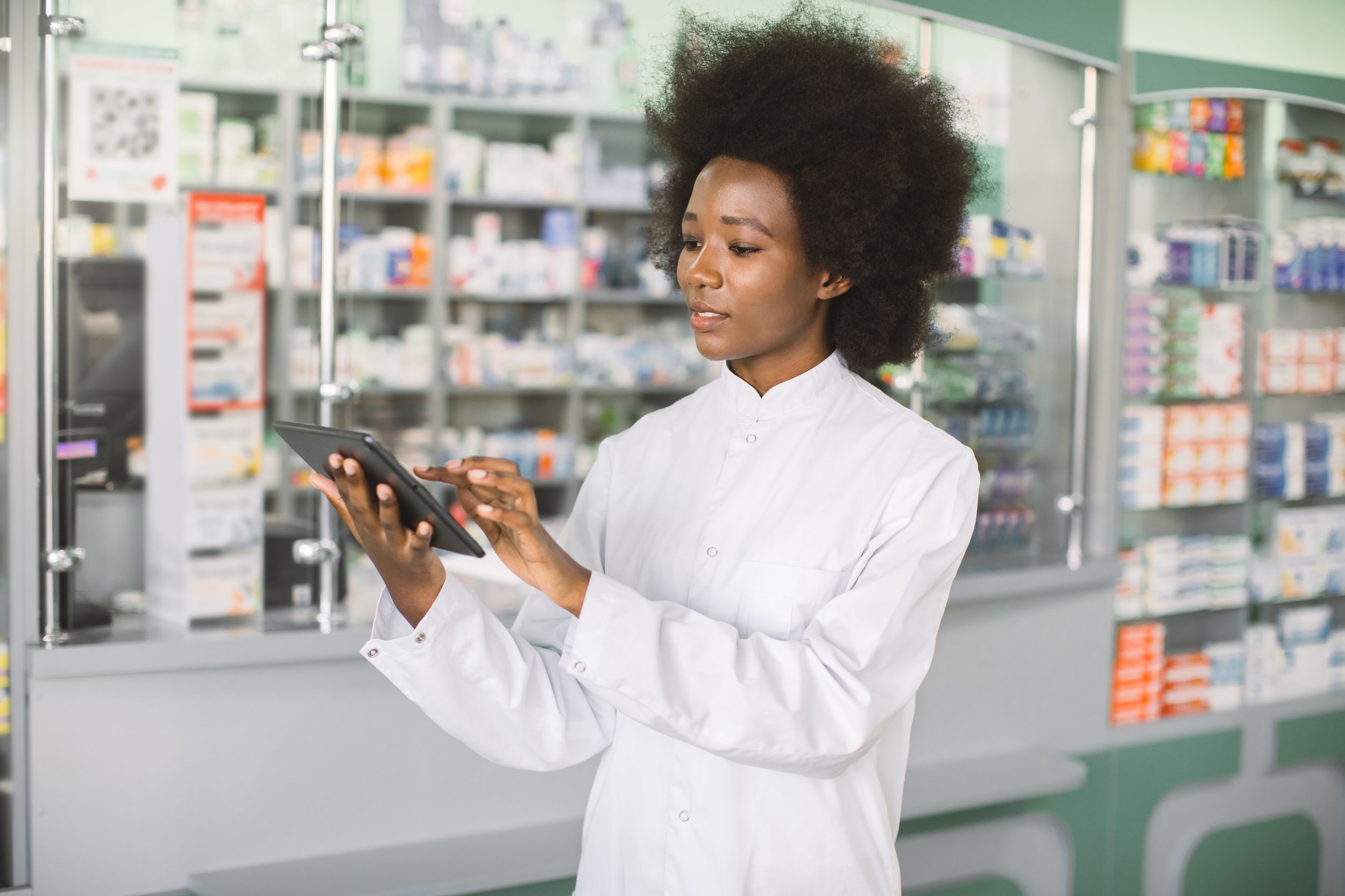 young confident pretty African woman doctor or pharmacist using digital tablet while standing in interior of modern hospital pharmacy or clinic. hospital pharmacy equipment